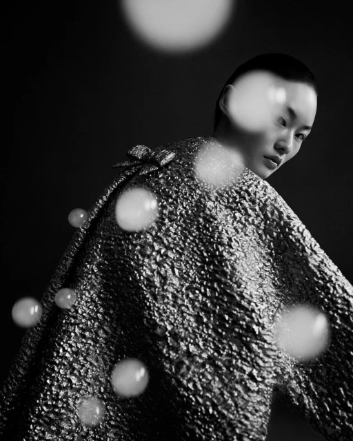 Monochrome portrait of a stylish Asian woman with short hair, posing in a reflective textured coat against a dark backdrop, studio lighting.