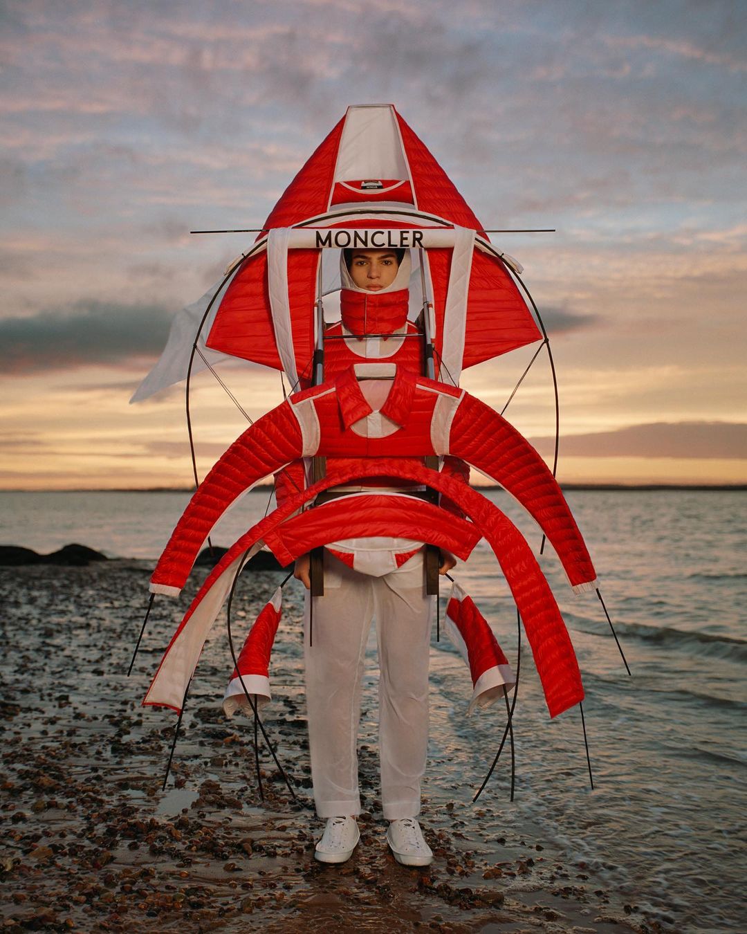 Fashion shot of a person wearing a red and white Moncler kite-like garment by the beach against a sunset sky.
