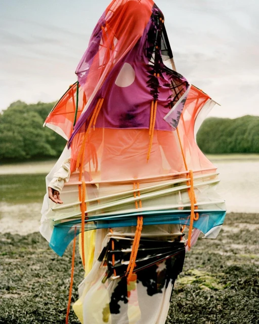 Person wearing colorful, patterned sheer fabric in an outdoor setting with water and trees in the background on an overcast day.