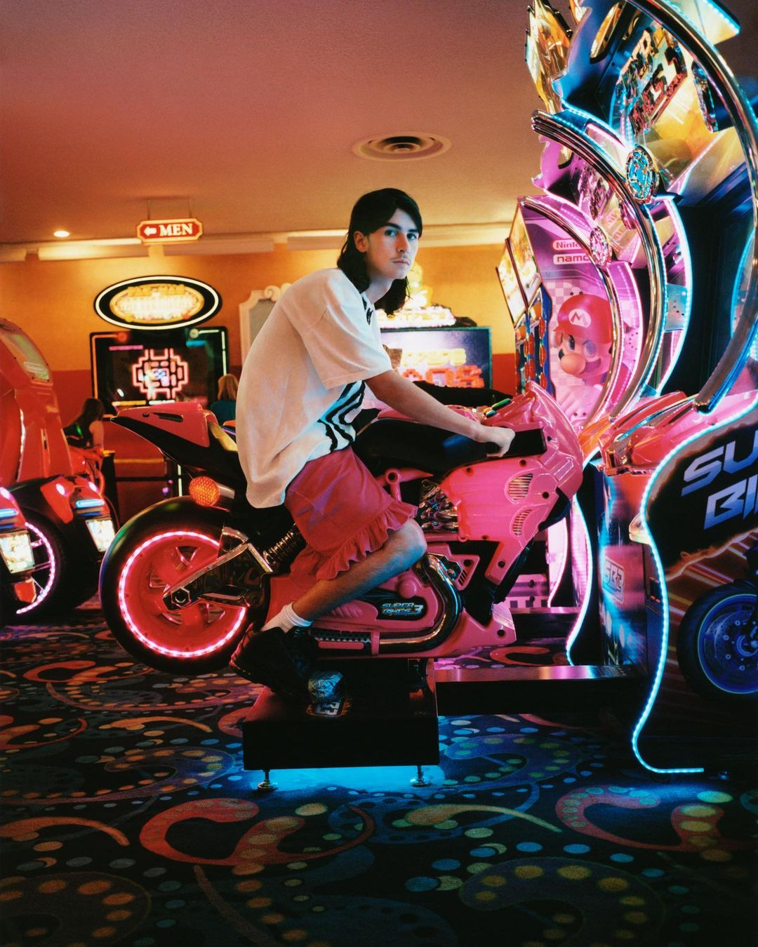 A person poses on a pink motorcycle arcade game inside an amusement arcade, with other games and carpeted flooring visible.
