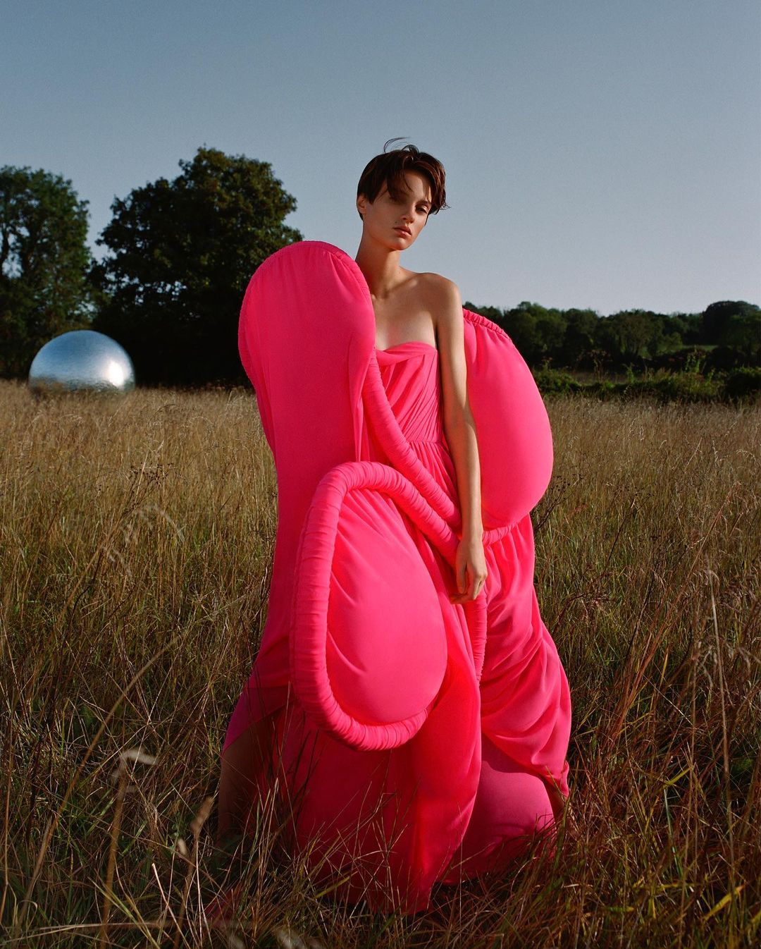 An adult female model in a pink gown standing in a field of tall grass, captured in a medium shot under a clear sky.