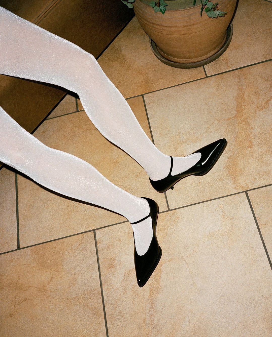 Elegant black Mary Jane heels on a light-skinned person wearing white tights, on a neutral-colored tiled surface with a potted plant.