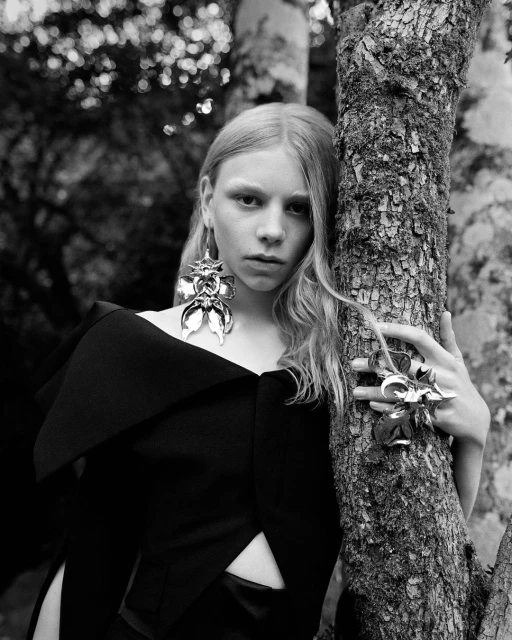 Black-and-white portrait of a young woman with blond hair and large, sculptural earrings, leaning against a tree in an outdoor setting.