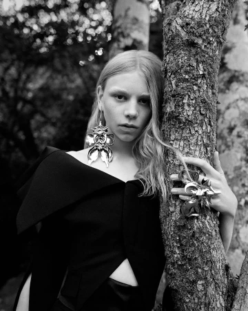 Black-and-white portrait of a young woman with blond hair and large, sculptural earrings, leaning against a tree in an outdoor setting.