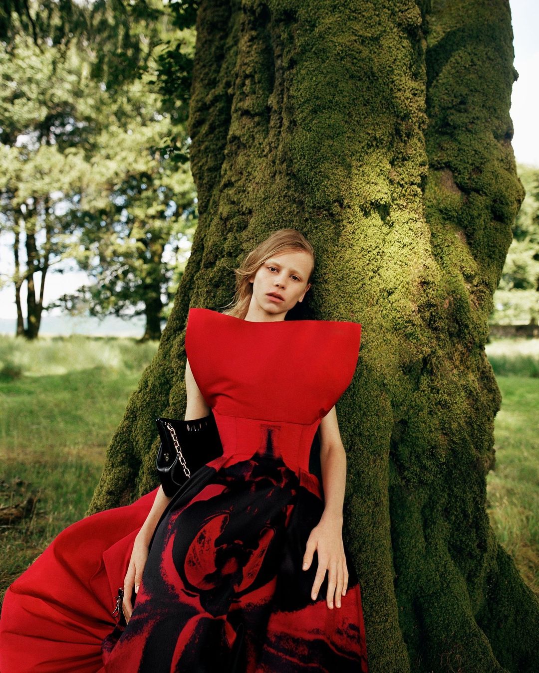 Young woman in red gown leans against a moss-covered tree in a forest setting. Fashion editorial with dramatic red dress.