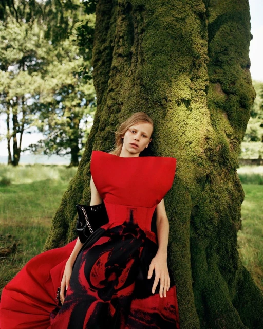 Young woman in red gown leans against a moss-covered tree in a forest setting. Fashion editorial with dramatic red dress.