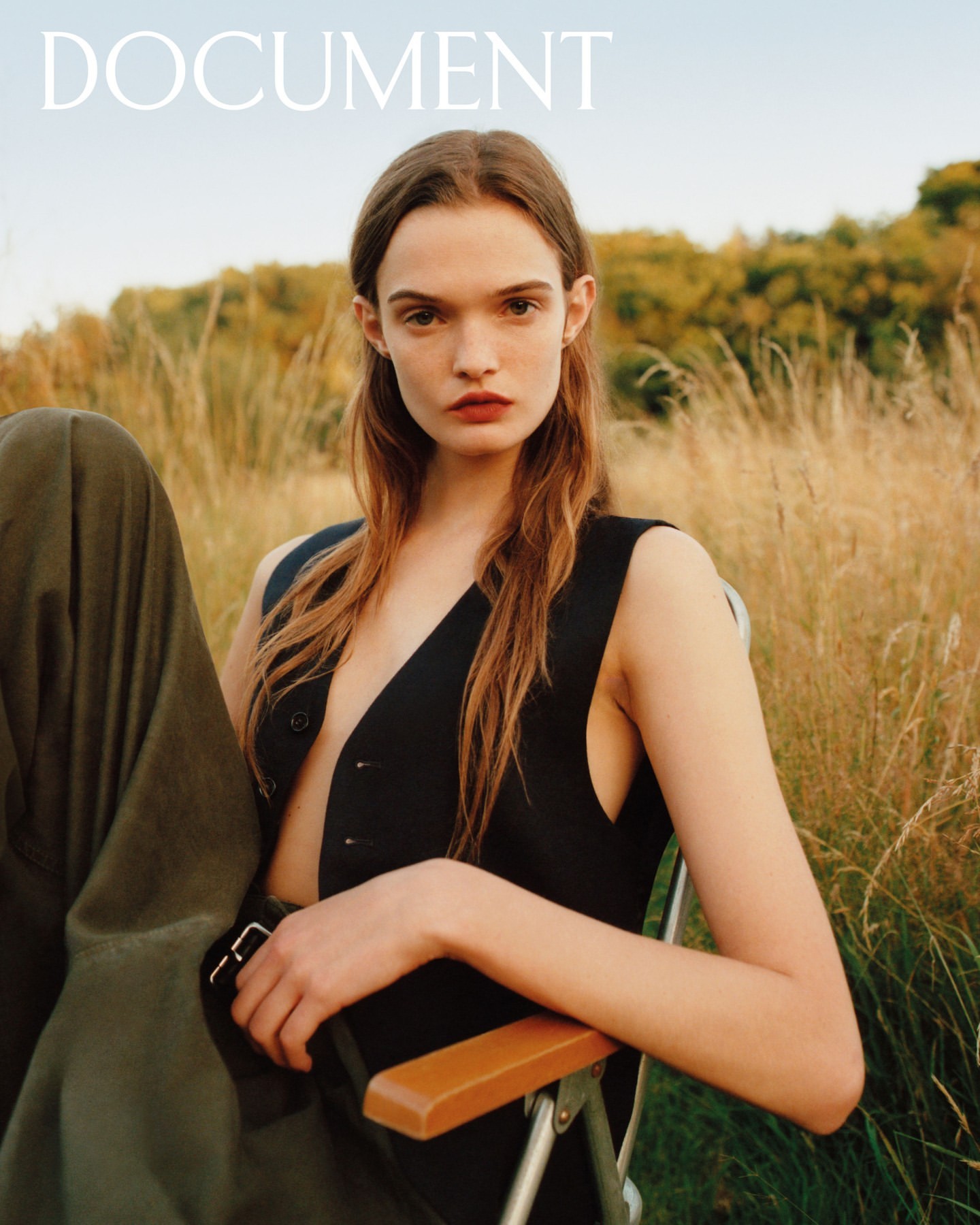 A young woman with auburn hair sits in a chair in a field of tall grass, wearing a black vest, in a full portrait with natural lighting.
