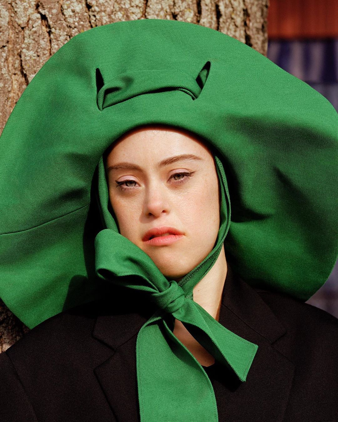 Close-up of a young woman with light skin and Down syndrome, wearing a black jacket and large green hat with a green bow. 