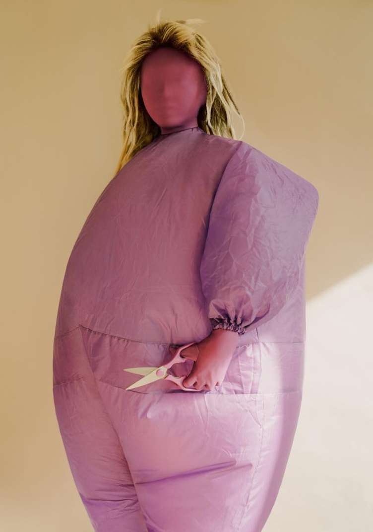 Blonde woman wearing a puffy lavender snowsuit with face covering, holding pink scissors in a studio with cream background.