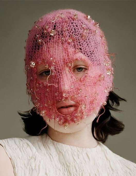 Close-up of a person wearing a decorative pink knit balaclava with small gems, fair skin, a neutral expression, and a white top.