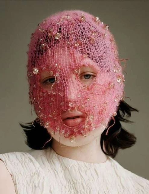 Close-up of a person wearing a decorative pink knit balaclava with small gems, fair skin, a neutral expression, and a white top.