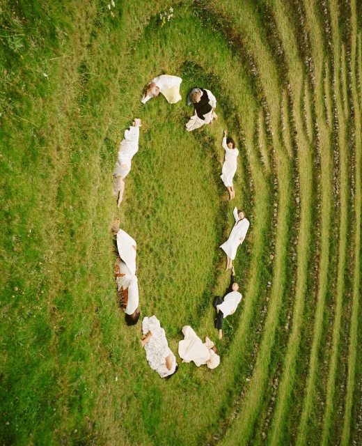 Aerial view of a group of people lying on a grassy field in a circle formation, creating a unique visual composition on the green landscape.
