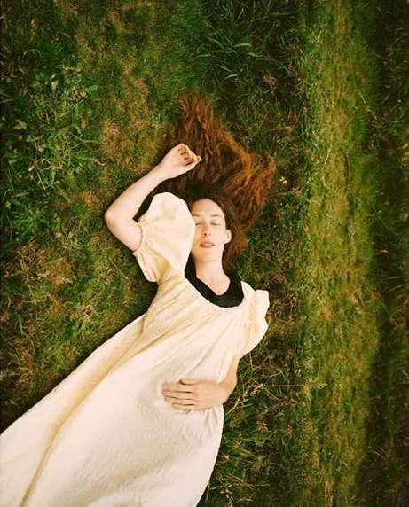 Woman with red hair lies on green grass wearing a light yellow dress. Relaxed pose in nature with soft, diffused lighting.