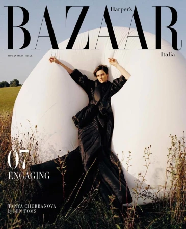 Model in black sculptural dress leaning against a large white inflatable sphere in an outdoor field with tall grass.