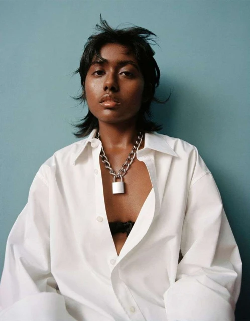 Portrait of a South Asian woman in an oversized white shirt and chunky padlock necklace against a muted teal backdrop.