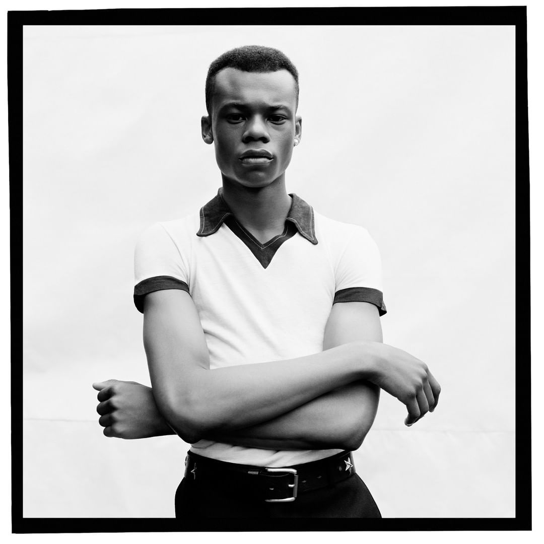Portrait of a young African American man with short hair, wearing a collared shirt and belt with star details, against a white backdrop.