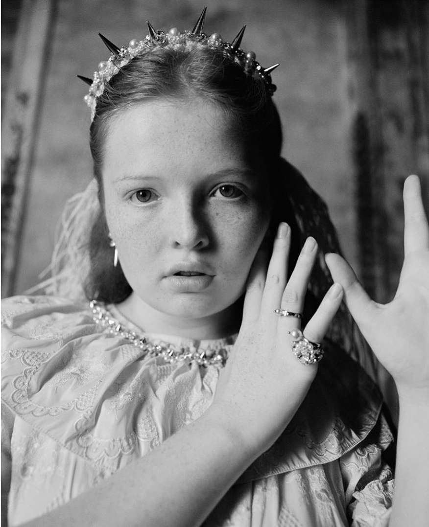 Portrait of young woman wearing spiked tiara and ruffled dress with hands raised by Ben Toms.