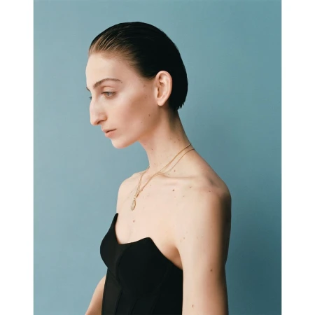 Profile portrait of a woman with slicked-back dark hair wearing a black strapless top and a gold pendant necklace against a blue background.