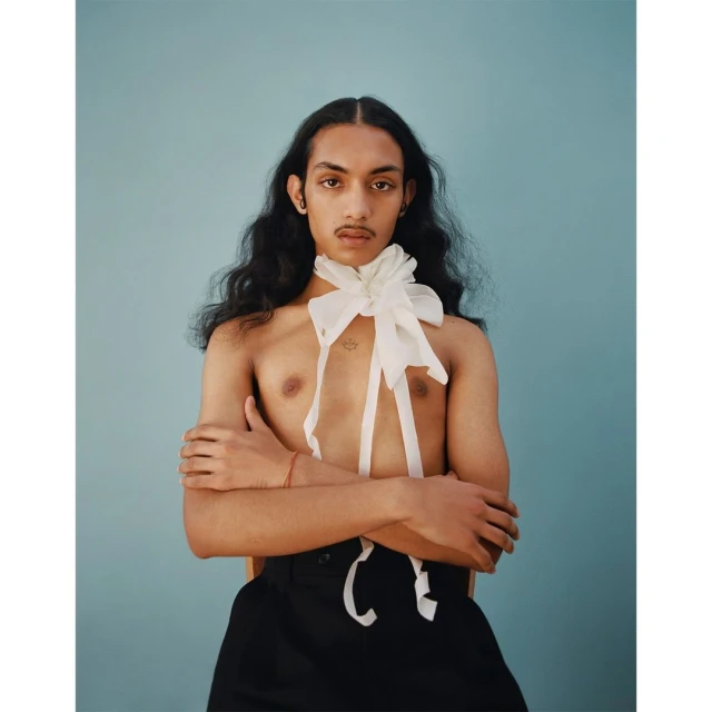 Portrait of a young adult person with long dark hair, wearing a large white bow and black pants, against a light blue background.