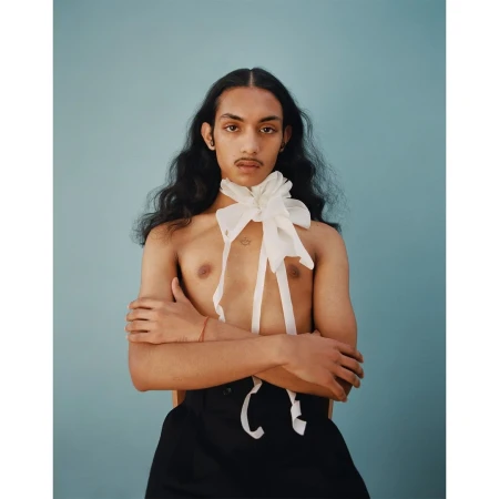 Portrait of a young adult person with long dark hair, wearing a large white bow and black pants, against a light blue background.