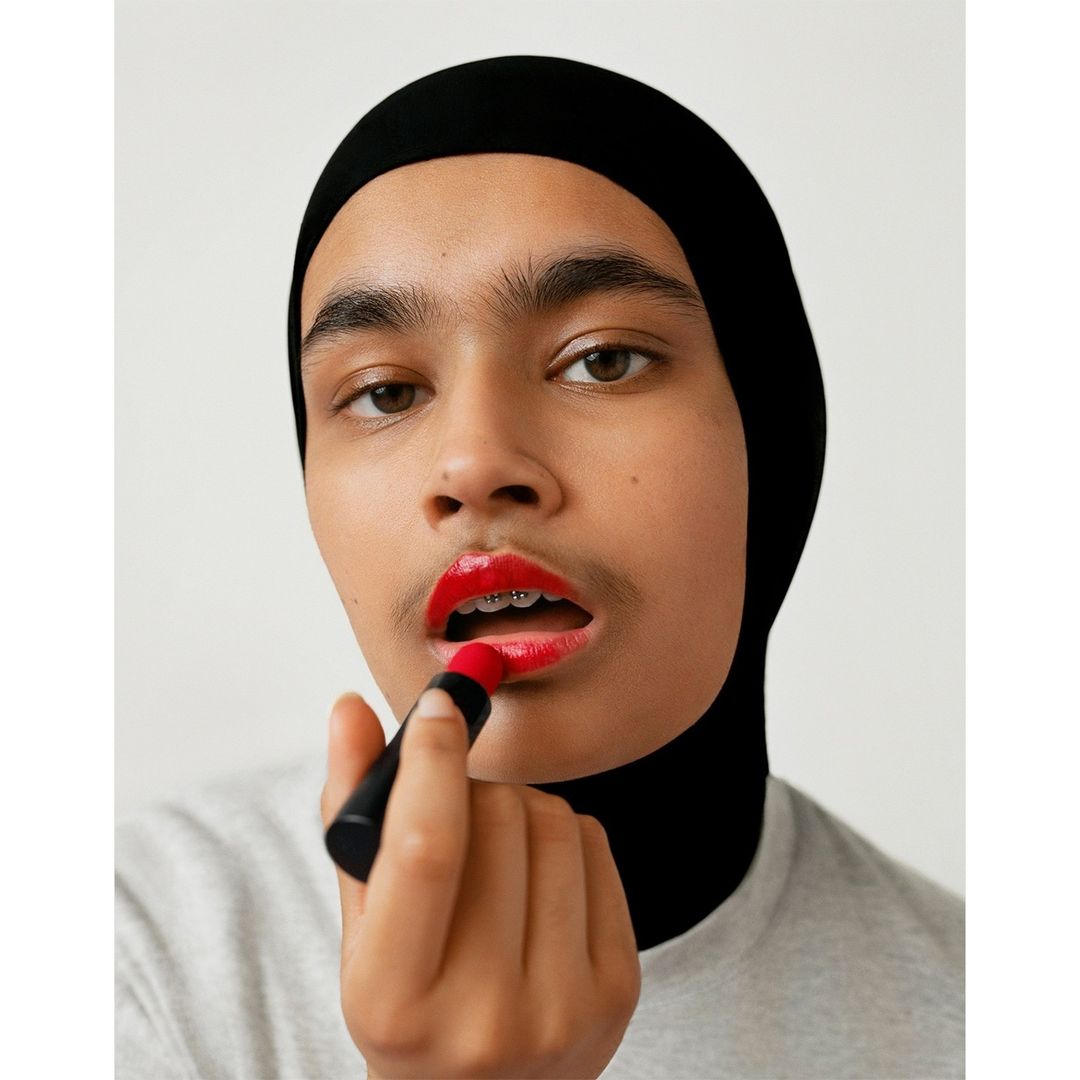 Close-up portrait of a young adult with a head covering applying bright red lipstick against a plain light background in a beauty setting.