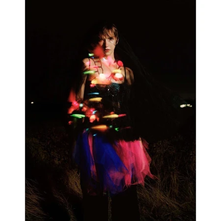 A female adult poses outdoors at night, wearing a pink and blue tutu and illuminated string lights wrapped around her neck in the darkness.