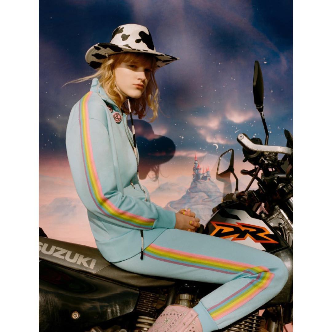 Adult female model in an athletic suit and cowboy hat poses with a motorcycle against a dreamy landscape backdrop with pastel colors.