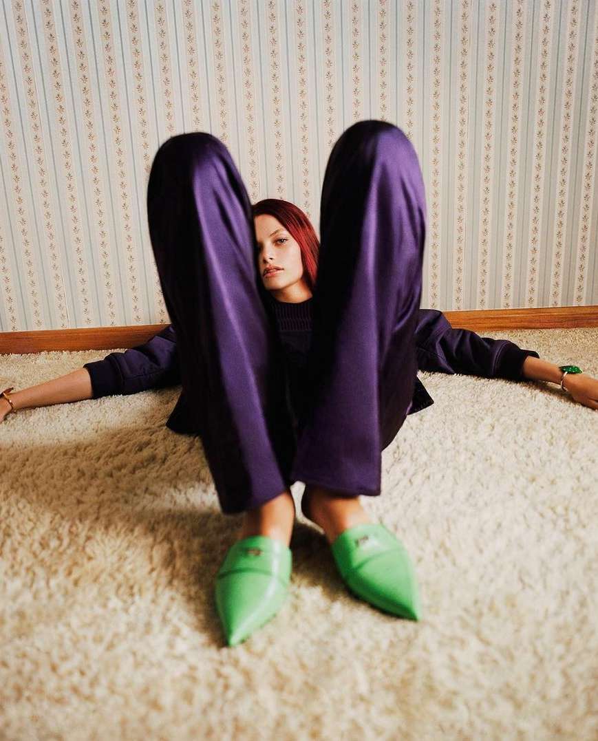 A woman reclines on a shag carpet in purple pants and green shoes in front of a floral wallpaper, shot with eye-level angle.