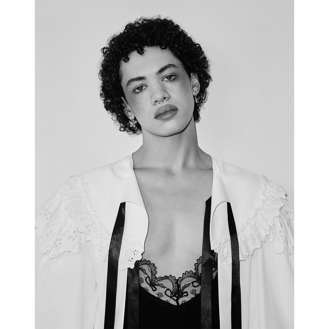 Black-and-white portrait of a person with curly hair wearing a lace-trimmed jacket and top, set against a plain light-grey background.