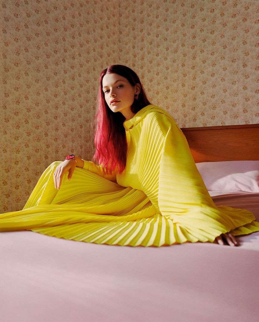 A woman with red hair sits on a pink bed in a yellow pleated dress. The room has floral wallpaper and soft light. Fashion editorial.
