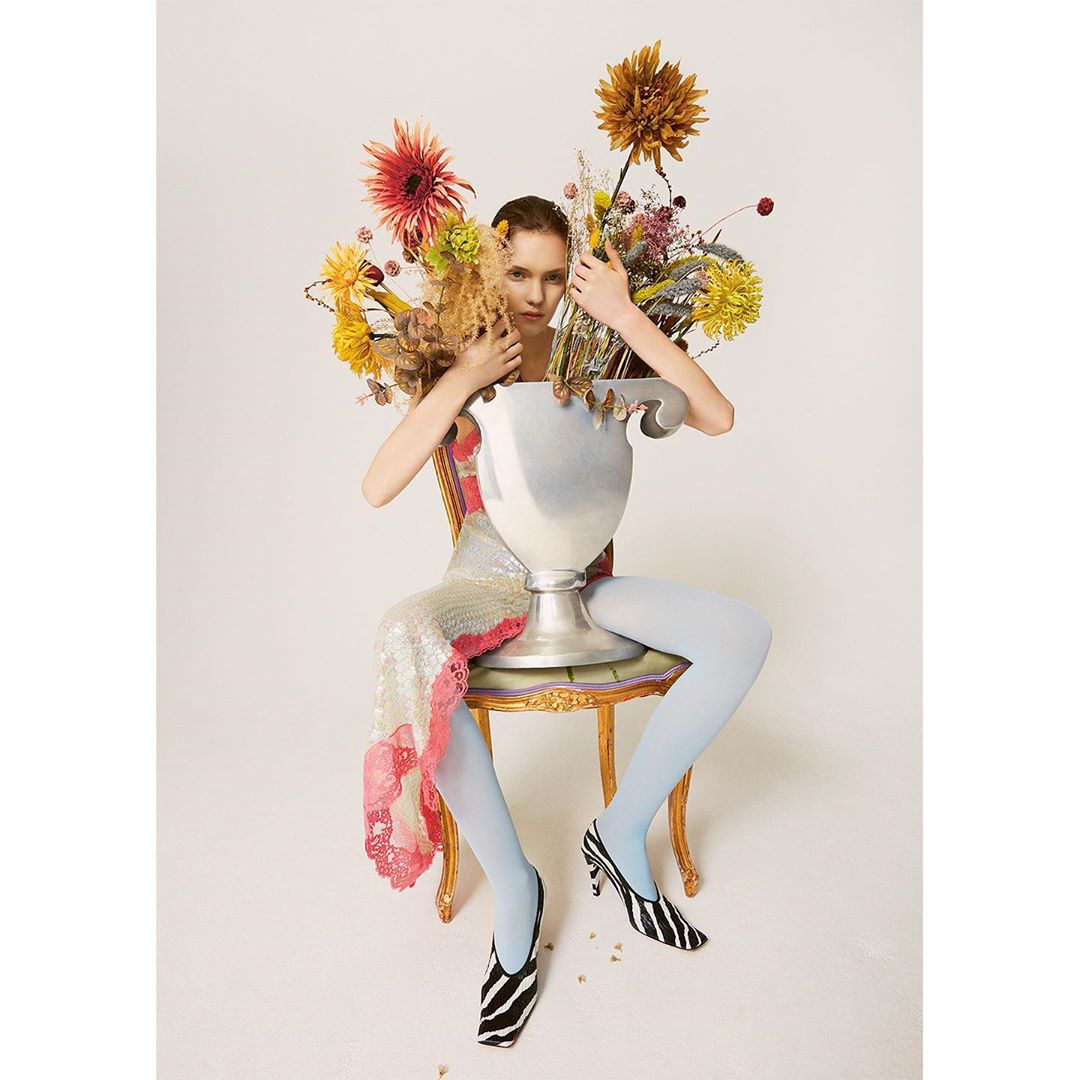 Woman in sequin dress sits on ornate chair behind a large silver vase filled with colorful flowers in a light, bright photo studio.