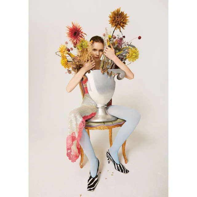Woman in sequin dress sits on ornate chair behind a large silver vase filled with colorful flowers in a light, bright photo studio.