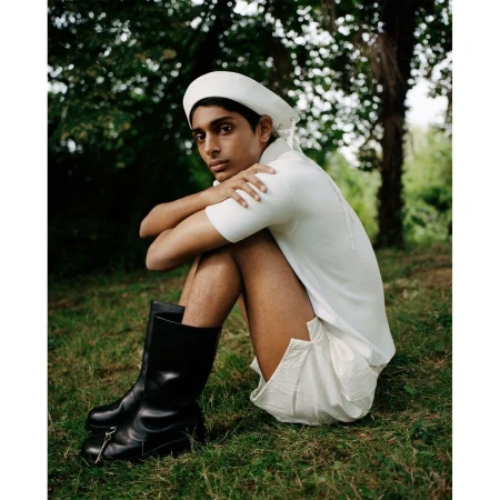 A young South Asian man wearing a white polo shirt, white shorts, white cap, and black boots sitting on grass outdoors.