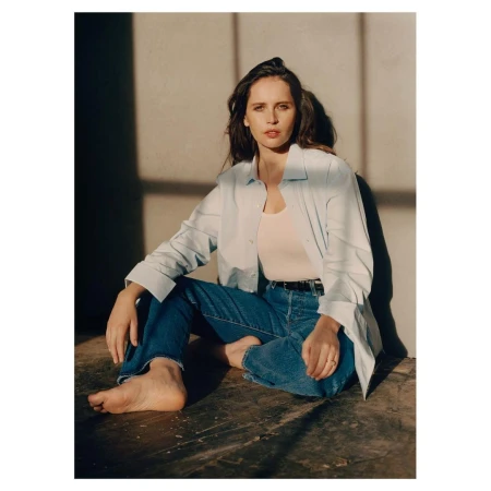 Felicity Jones seated on the floor wearing an unbuttoned white shirt, cream top, and blue jeans, lit by strong sunlight casting window shadows on the wall.