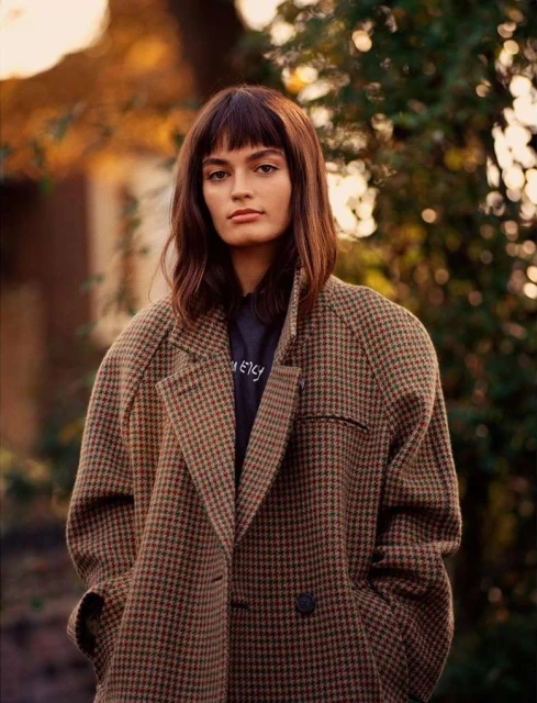 A young woman with bangs in a patterned brown coat stands outdoors against a natural backdrop with soft, warm lighting. Fashion shot.