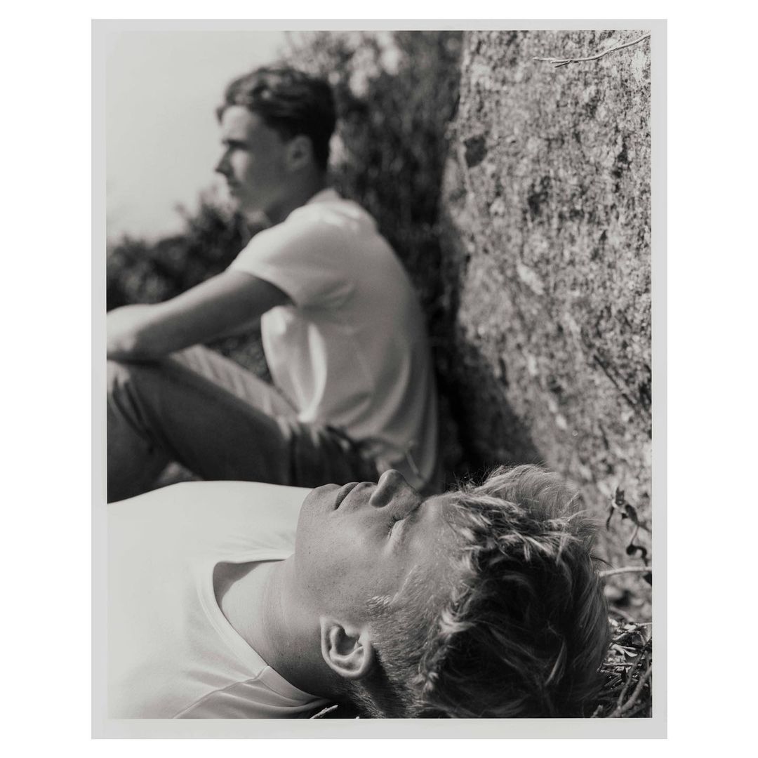 Black and white portrait of two young adult males outside, one reclining with eyes closed, the other sitting up looking away.