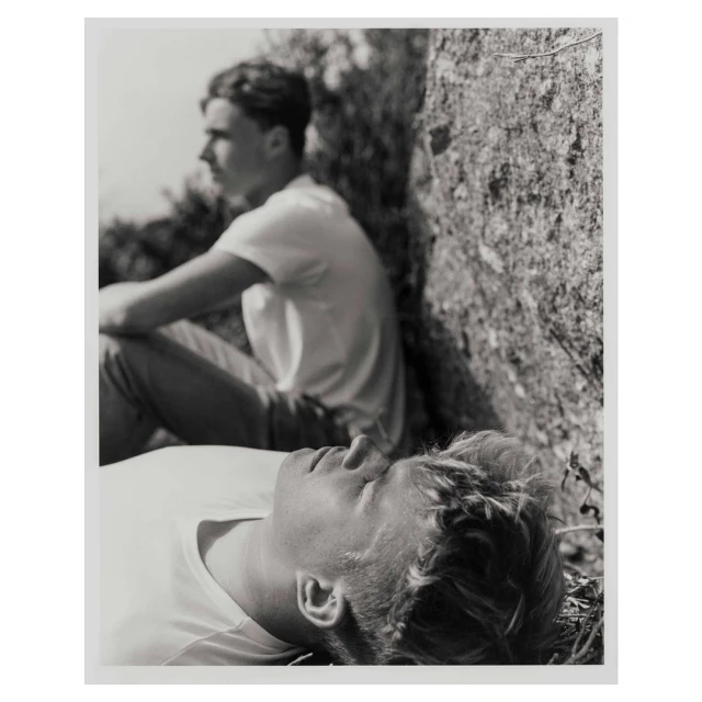 Black and white portrait of two young adult males outside, one reclining with eyes closed, the other sitting up looking away.