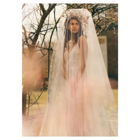 Woman in tulle dress and floral headpiece stands in front of tree branches and an out-of-focus building, in a dreamy, outdoor setting.