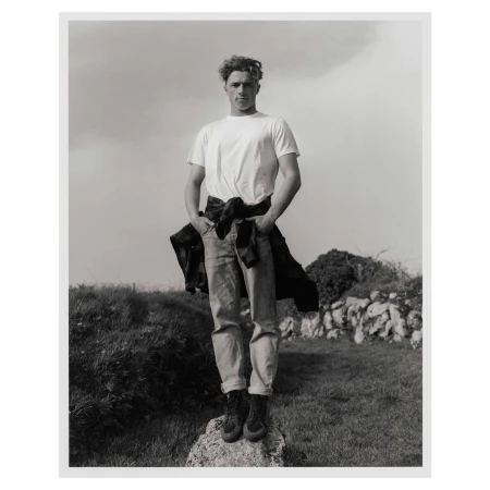 Black and white portrait of a young man standing on a rock wearing a white t-shirt, jeans, and a jacket tied around his waist.