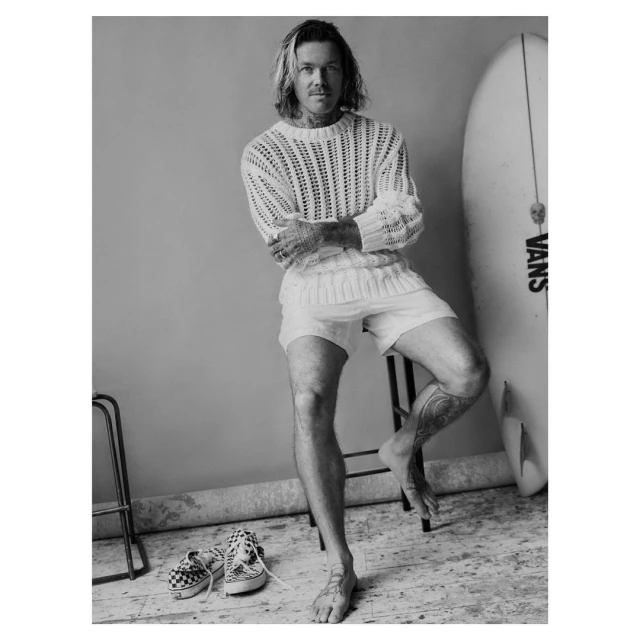 Monochrome portrait of a man with long hair sitting on a stool, wearing a knit sweater and shorts, with a surfboard and shoes.
