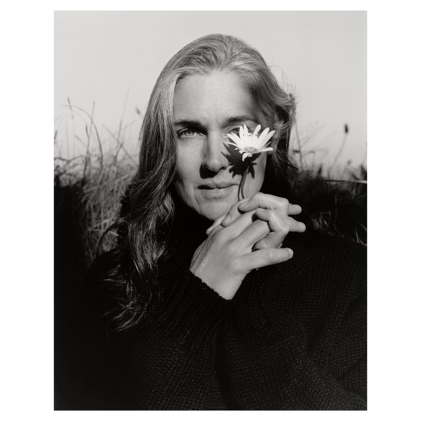 Black and white portrait of an adult woman with long grey hair, posing outdoors with a white daisy flower near her eye.