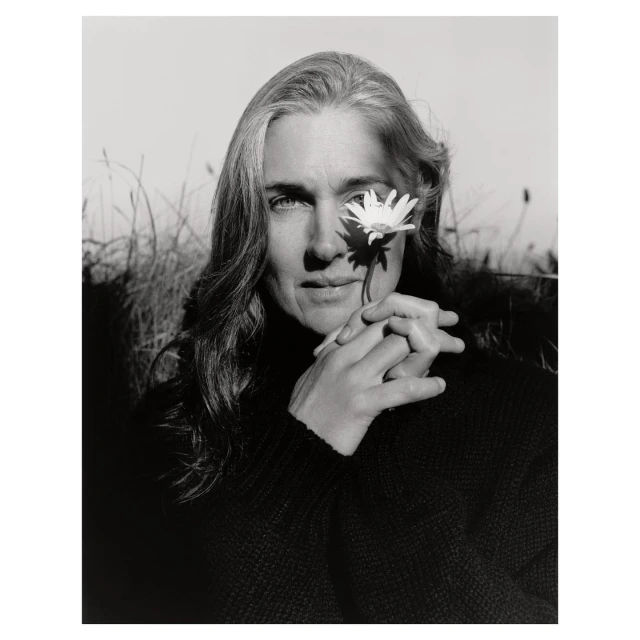 Black and white portrait of an adult woman with long grey hair, posing outdoors with a white daisy flower near her eye.
