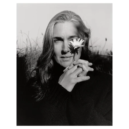 Black and white portrait of an adult woman with long grey hair, posing outdoors with a white daisy flower near her eye.