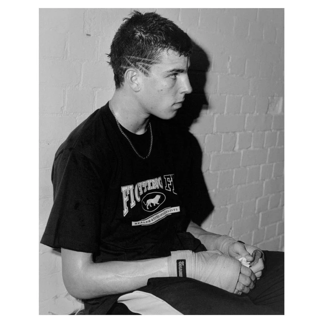 Black and white portrait of a seated boxer wearing a t-shirt with wrapped hands, photographed indoors against a brick wall background.