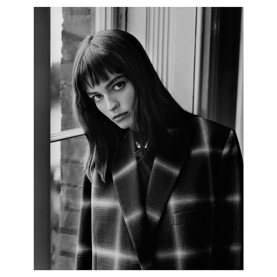 Portrait of a woman with bangs wearing a dark plaid blazer standing indoors near a window, captured by Ben Weller.