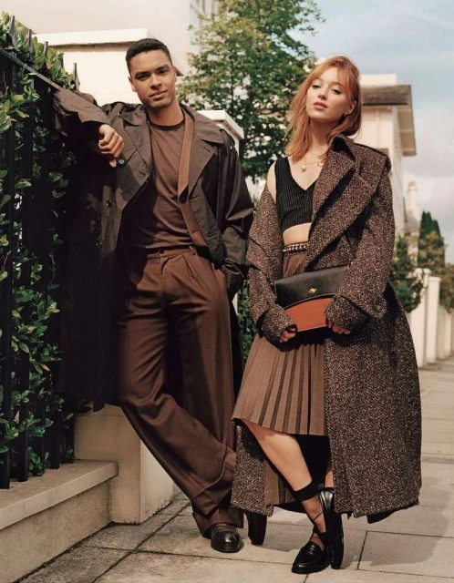 Two fashion models strike a pose outdoors, showcasing stylish brown outfits against a backdrop of greenery and buildings on a sunny day.