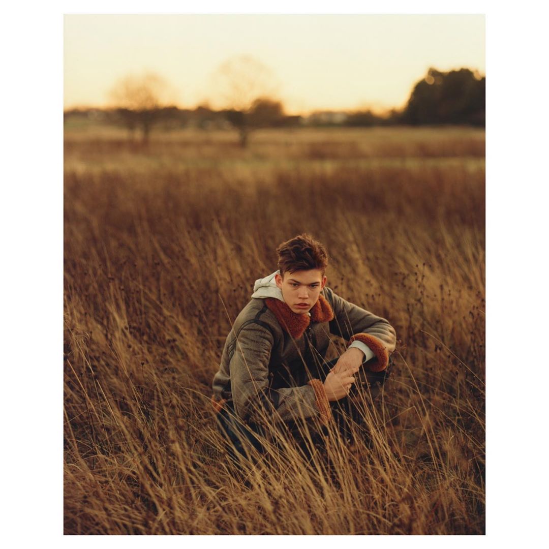Adult male sits in a grassy field wearing a fur-lined jacket at dusk with trees in the background in a warm, outdoor fashion shot.