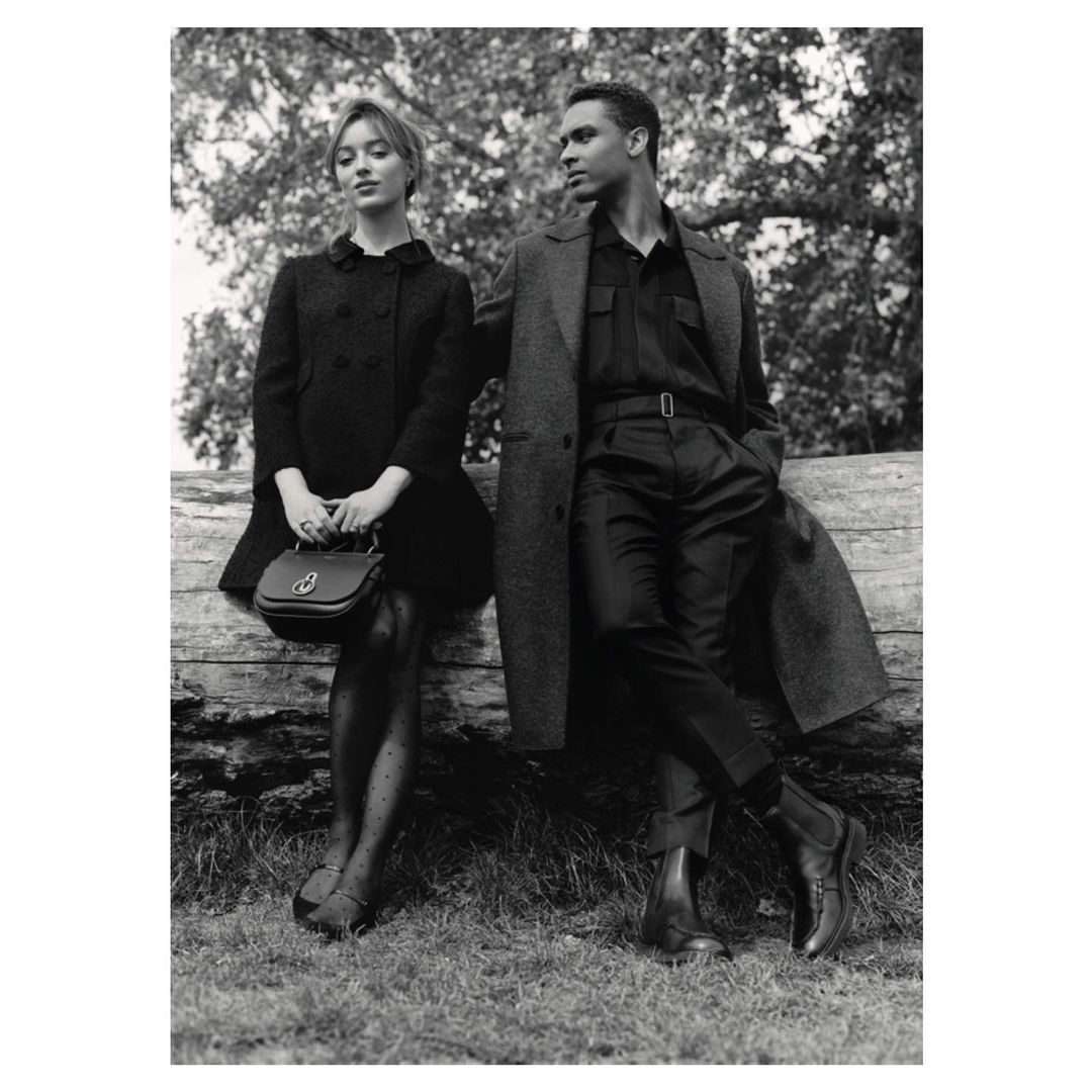 Black and white fashion shot of an adult couple posing outdoors, leaning against a fallen tree in a park setting, wearing dress clothes.