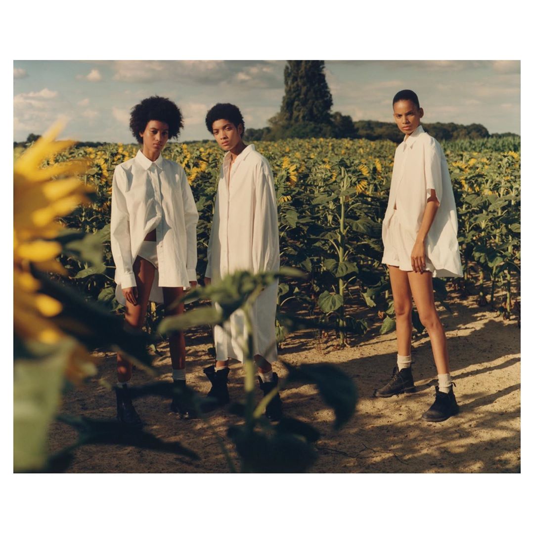 Three fashion models in white shirts and dark boots pose in a sunflower field under a partly cloudy sky, capturing a blend of nature and style.