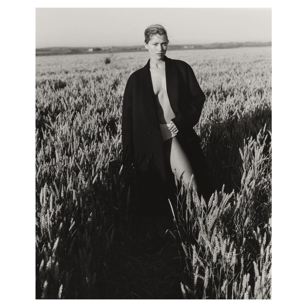 Black and white portrait of a woman in a coat standing in a wheat field, monochromatic and textural, with a rural outdoor setting.